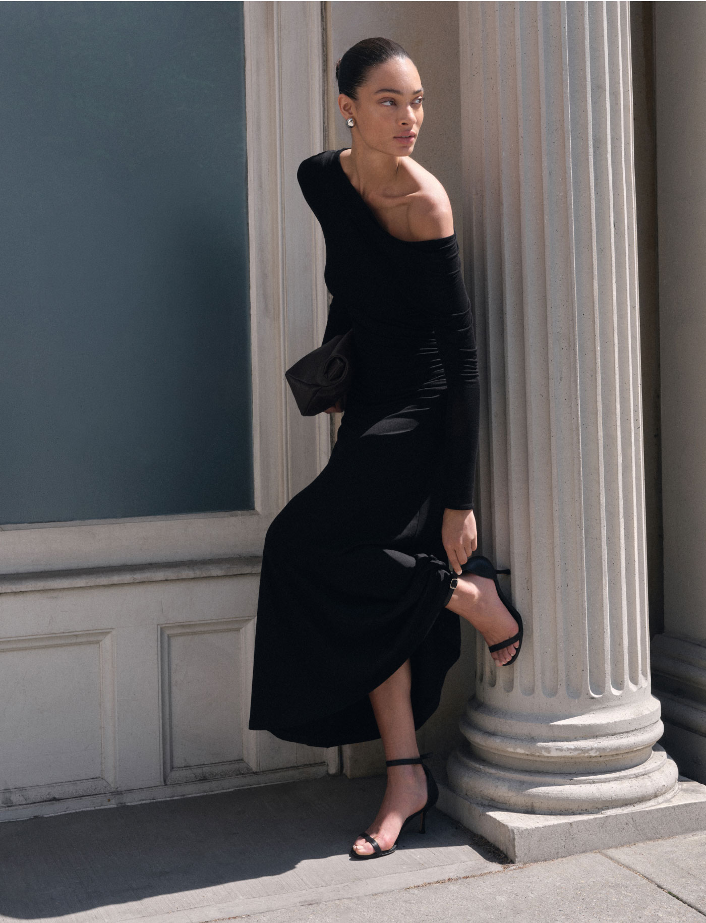 Aliza poses beside a classical column in a long black off-the-shoulder Ambrose Dress with black strappy heels and a black clutch.
