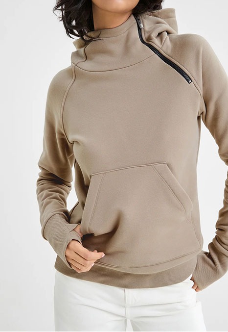 Emma wears a sand Booth Sweatshirt with an asymmetric zip collar, kangaroo pocket, ribbed cuffs, and white pants.