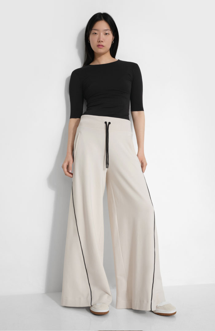 Xun wears a cream Seneca Pant with a drawstring waist and contrasting black piping along the legs, styled with a fitted black top and white shoes.