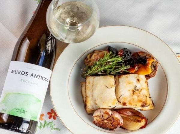 Bottle and glass of Muros Antigos Escolha Vinho Verde 2020 on a table next to a plate of fish and vegatables.