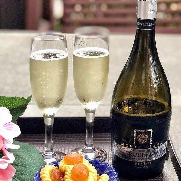 Chilled bottle of Prosecco Doc Treviso Extra Dry by Cantina Montelliana on a table with two glasses of wine with flowers and pastry.