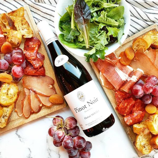Bottle of Pinot Noir 2020 by Florian Mollet lying label up on a table next to plates with dried meat, grapes, salad and bread