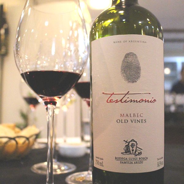 Bottle of Testimonio Old Vines Malbec 2019 next to several glasses of red wine placed on a table. 