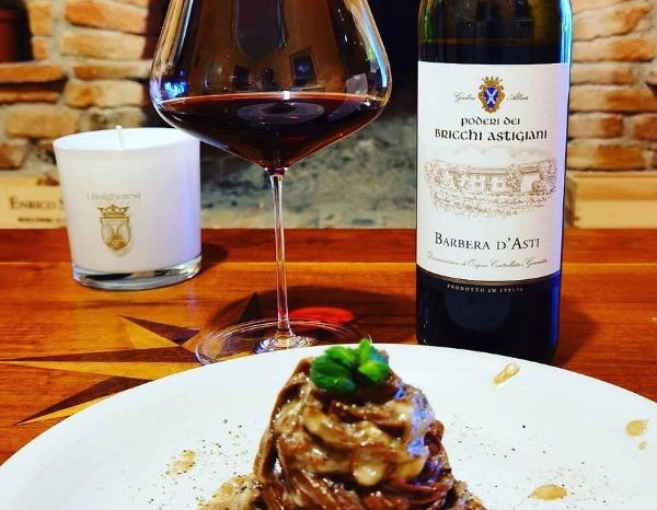 A bottle of Barbera D'Asti DOCG By Poderi Dei Bricchi Astigiani 2019 on a wooden table next to a glass of wine behind a plate of succulent pasta.