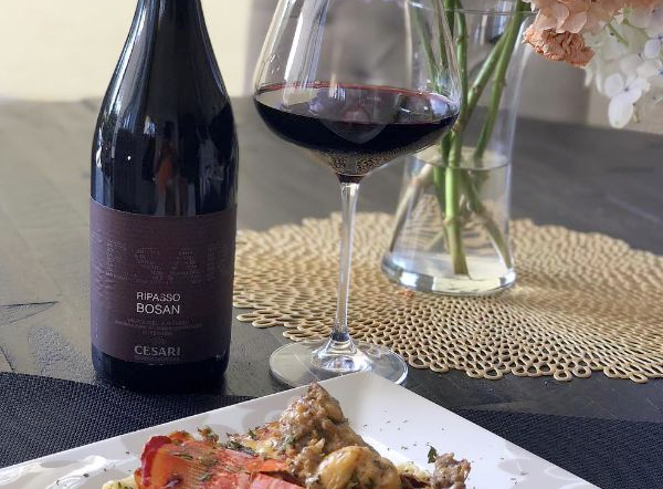 Bottle of Bosan Ripasso Valpolicella Superiore by Cesari 2017 next to a glass of wine on a table next to a vase of flowers and a plate of food.