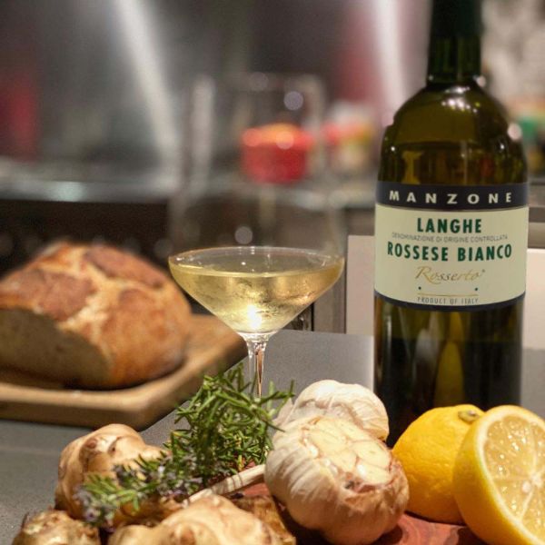 Bottle and glass of Langhe Rossese Bianco Rosserto 2018 on a table next to a loaf of bread, two half lemons, garlic bulbs and a sprig of rosemary.