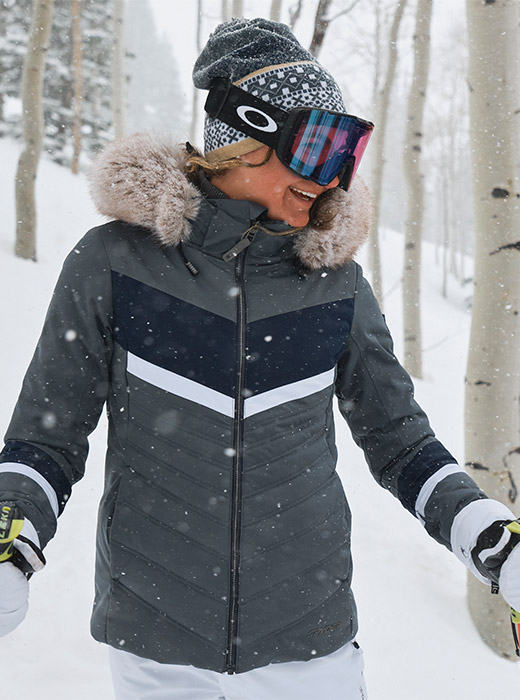 Female skier wearing a slate blue ski jacket with faux fur trim in a snowy forest.