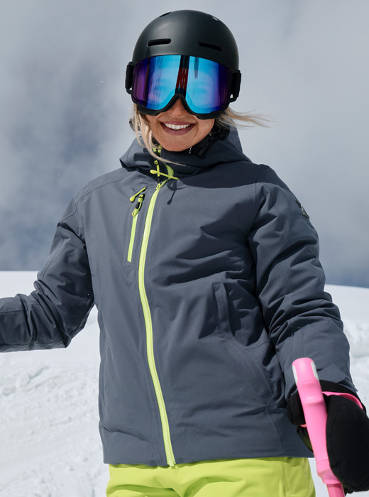 Female skier standing on a snow mountain top wearing a slate blue jacket with lime accents.