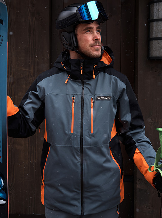 Skier standing with a wood wall backdrop holding skis.