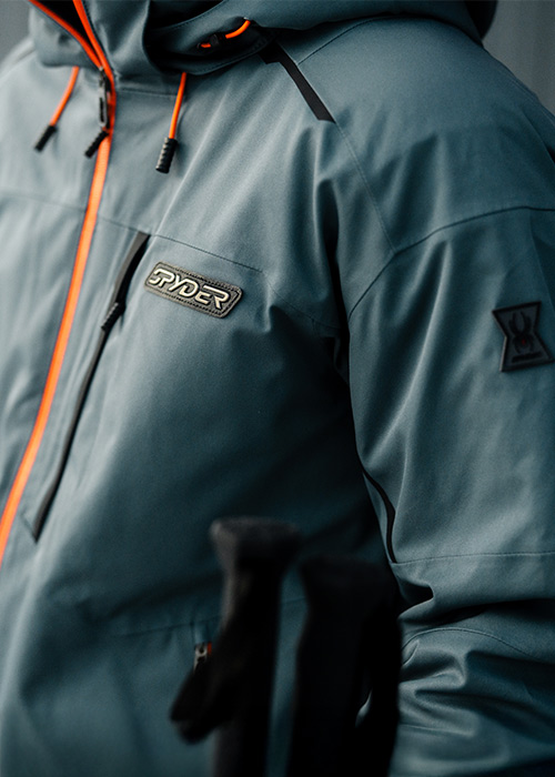 A close-up of a person wearing a slate blue Spyder jacket with orange accents, highlighting the logo and zipper details.
