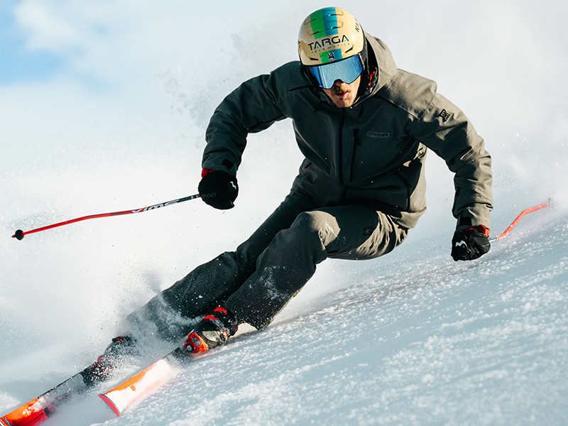 A skier in a grey suit expertly carves through fresh snow, sending up a spray of powder against a bright winter backdrop.