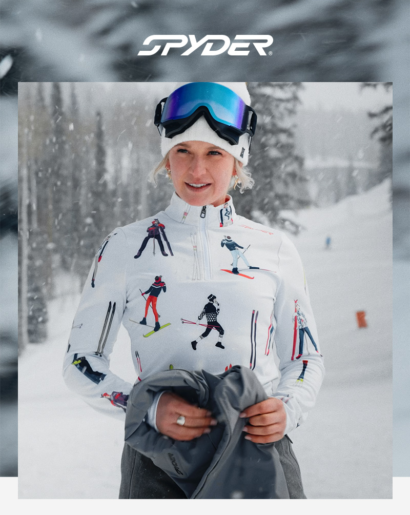 A skier smiles in a snowy landscape, wearing a colorful top and holding a jacket.