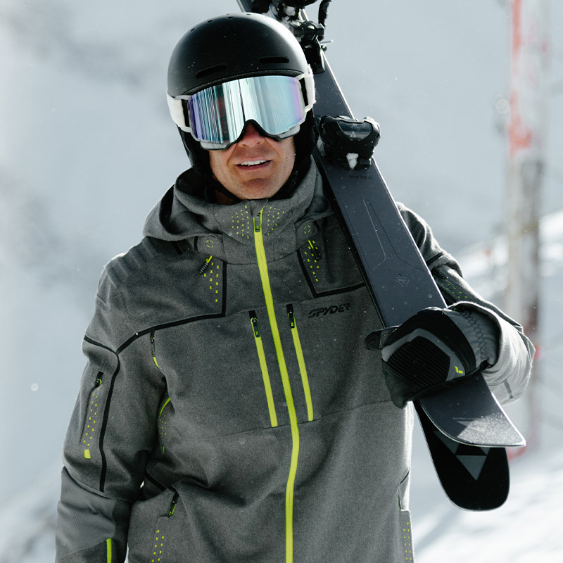 Man in sleek gray ski jacket with neon accents carries skis on shoulder.