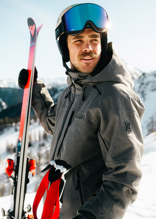 A skier in a gray jacket holds a pair of skis, set against a snowy mountain landscape under clear blue skies.