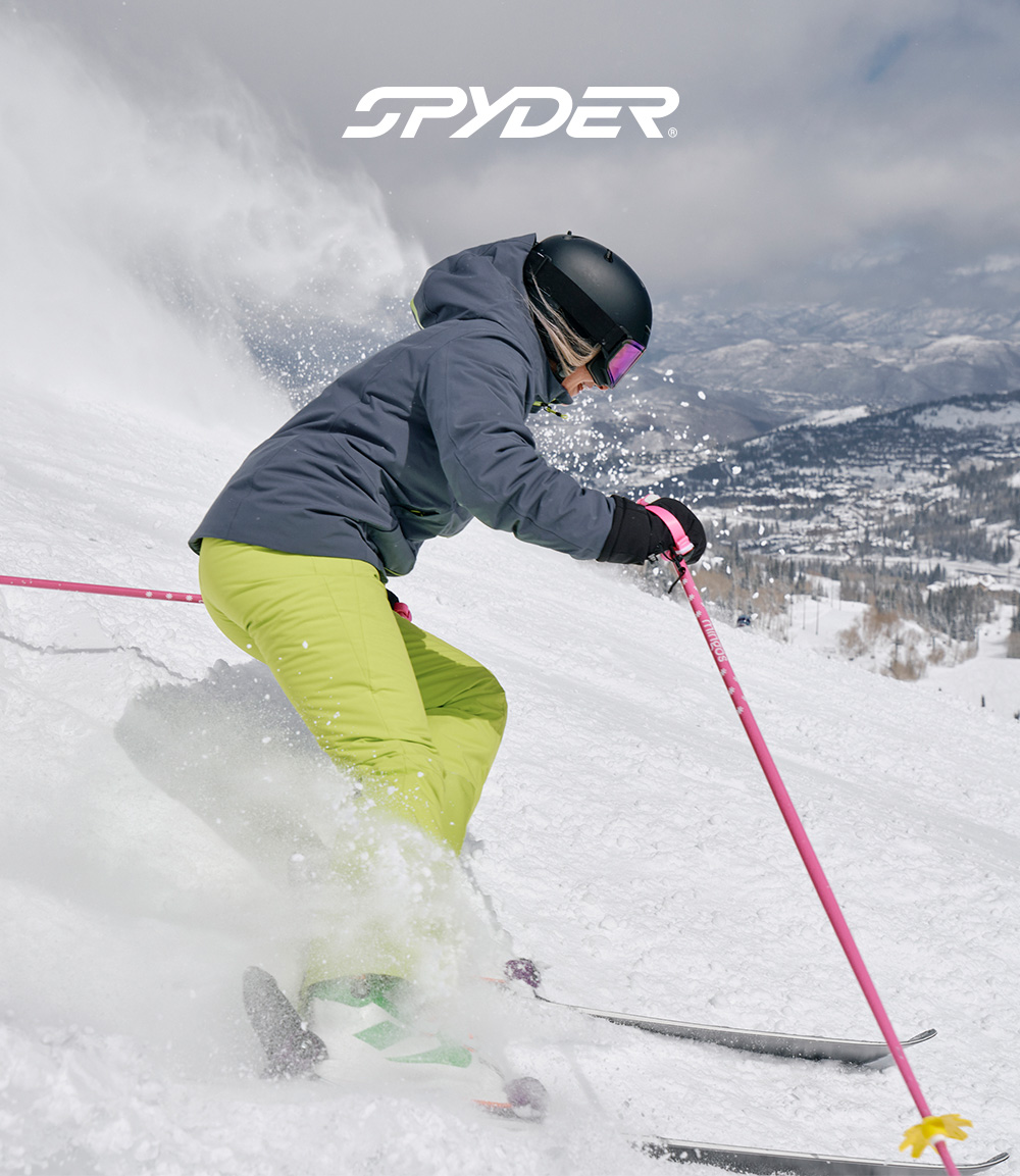 Skier going down the slide of a hill, wearing a slate blue ski jacket and lime pants.