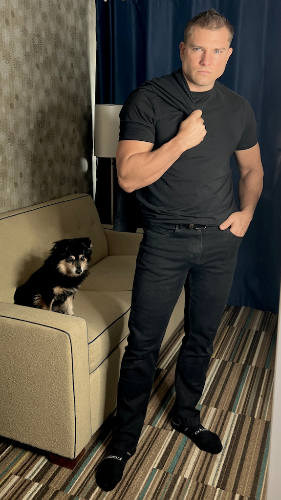 Man wearing a Black Crew Neck and Slim Bandits with a dog on the couch behind him