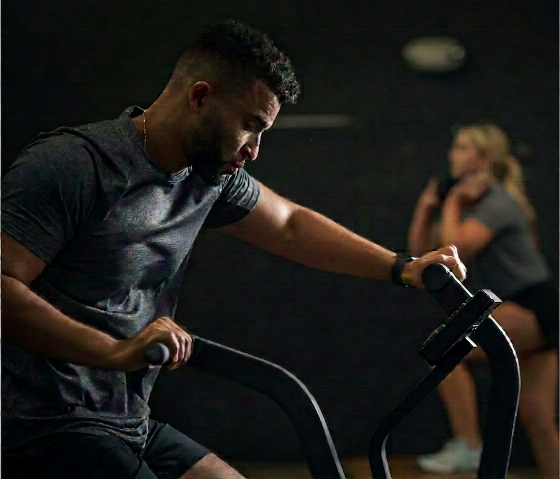 Man on an air bike with a woman doing squats in the background.
