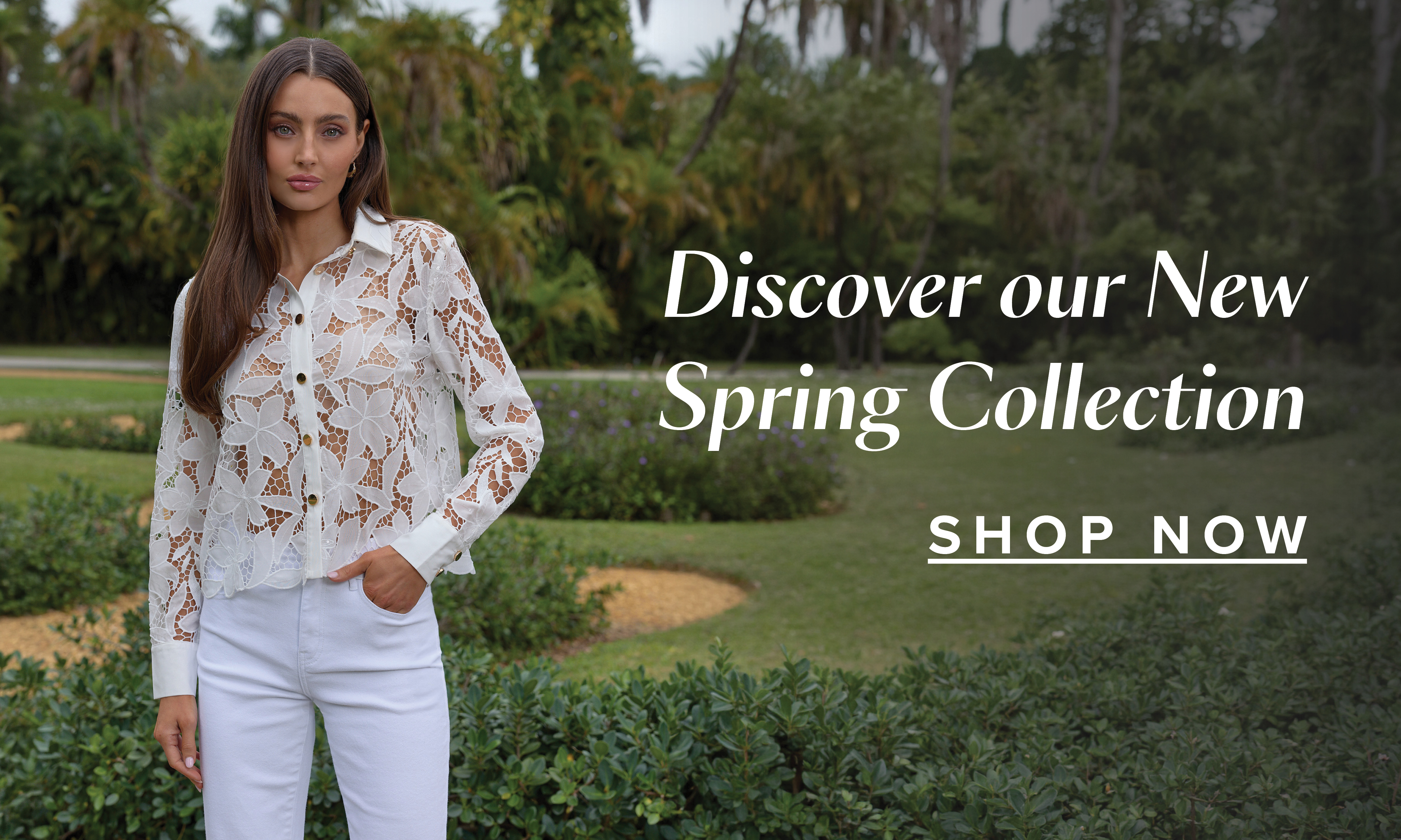 Discover Our New Spring Collection. Shop Now. A model standing posed wearing the White Mina Lace Blouse with a lush green garden behind her.