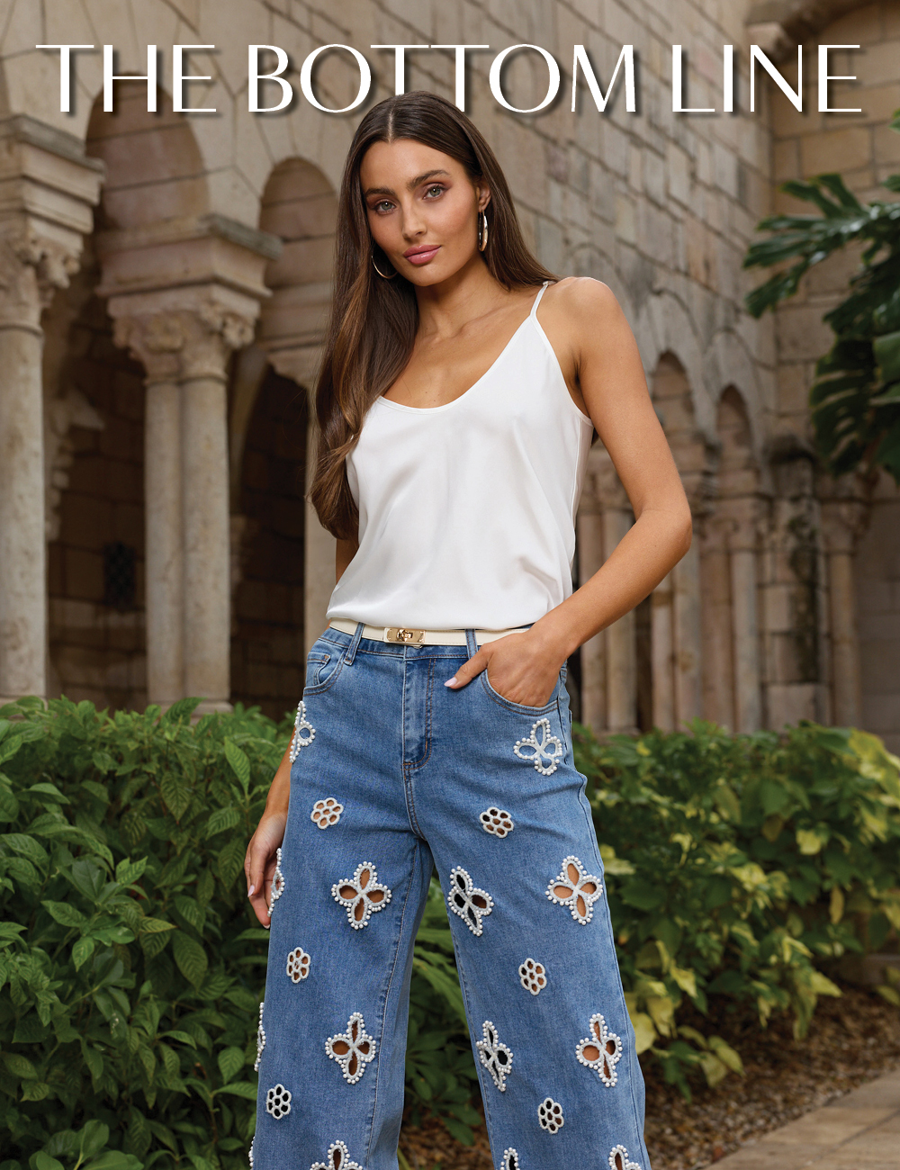 The Bottom Line. A model standing posed wearing the Pearl Yara Silk Camisole, Medium Blue Kaia Pearl High Rise Jean, and Pearl Nicola Belt with a lush garden and stone archways behind her.