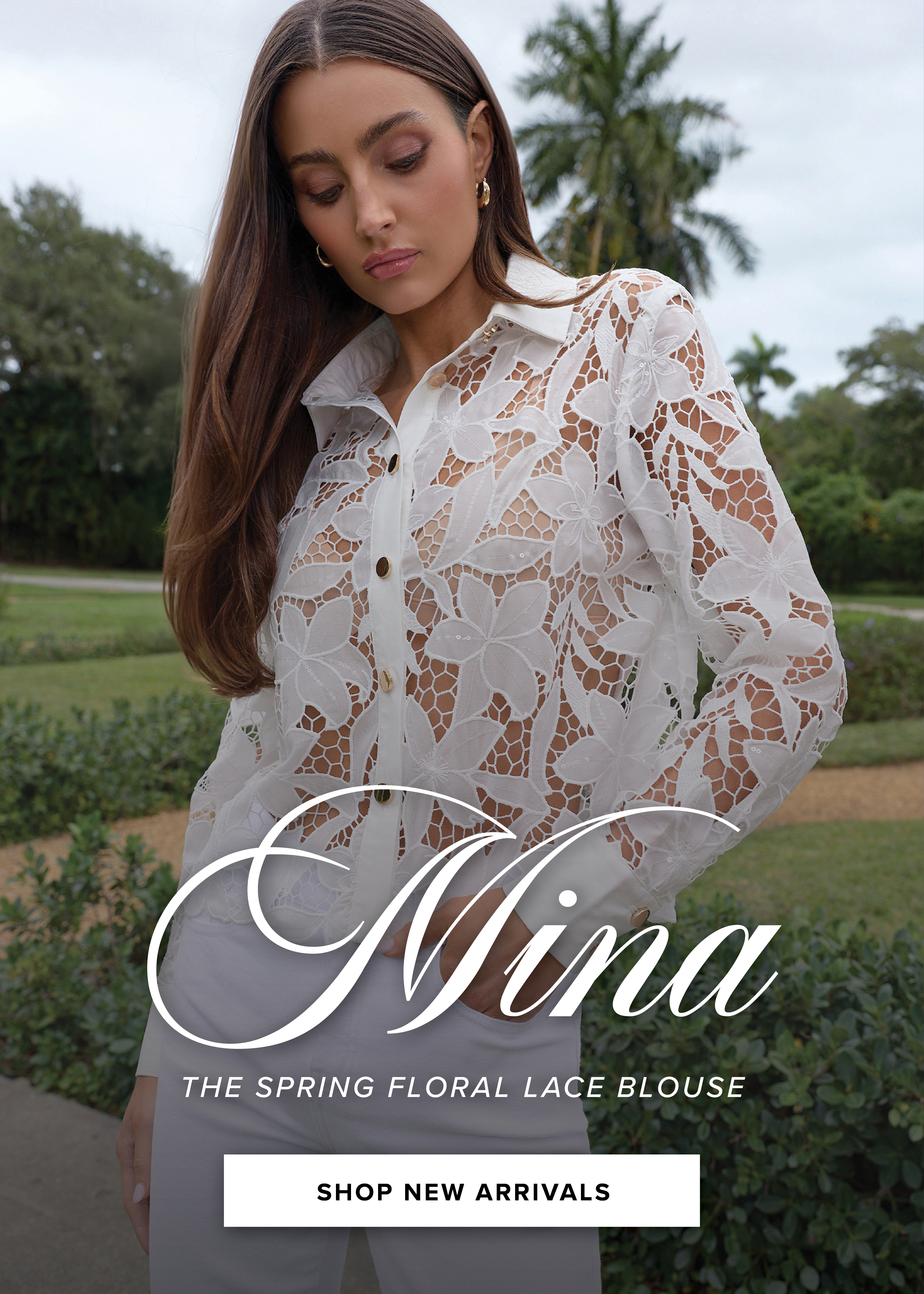 Mina. The Spring Floral Lace Blouse. Shop New Arrivals. A model standing posed close up wearing the White Mina Lace Blouse with a lush green garden behind her.