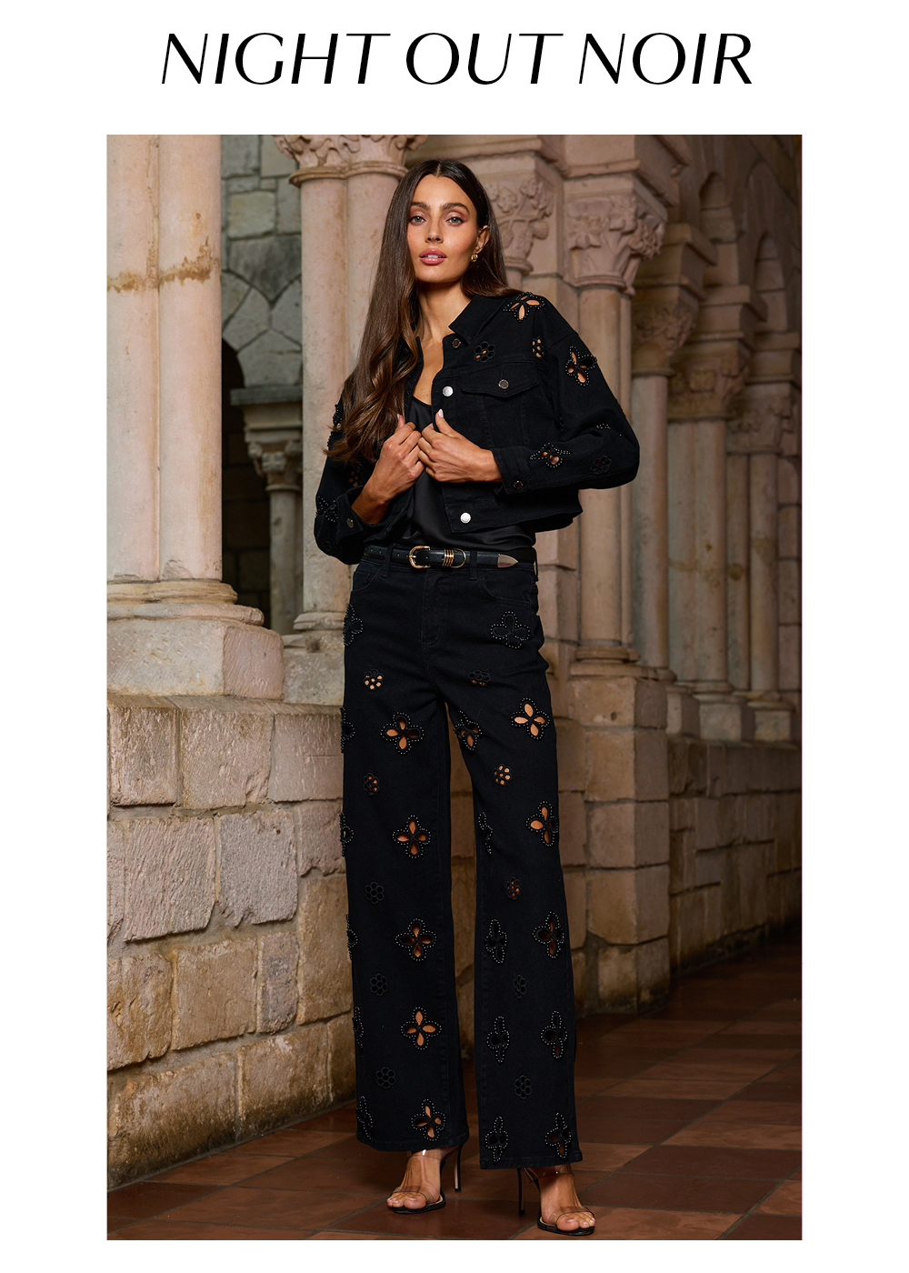 Night Out Noir. An image of a model in the Black Kaia Pearl Jean Jacket, Black Yara Silk Camisole, Black Kaia Pearl High Rise Jean, and Black Blair Belt posed in a stone corridor. 