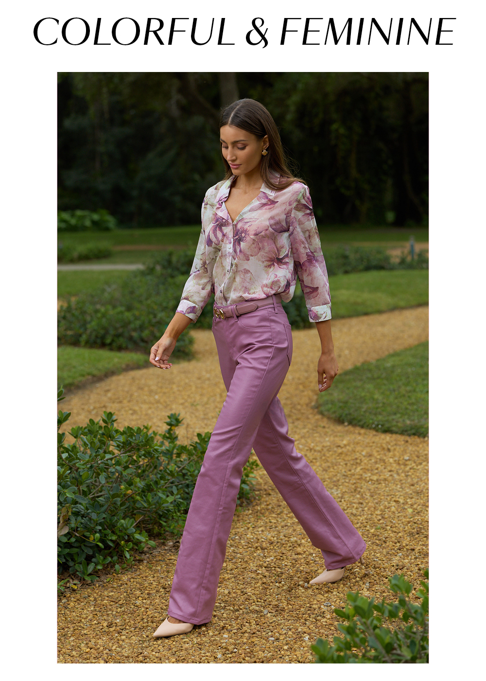Colorful and Feminine. An image of a model in French Garden Sara Chiffon Blouse, Mauve Aria Coated Flare Jean, and Bettie Belt walking in a lush green garden.
