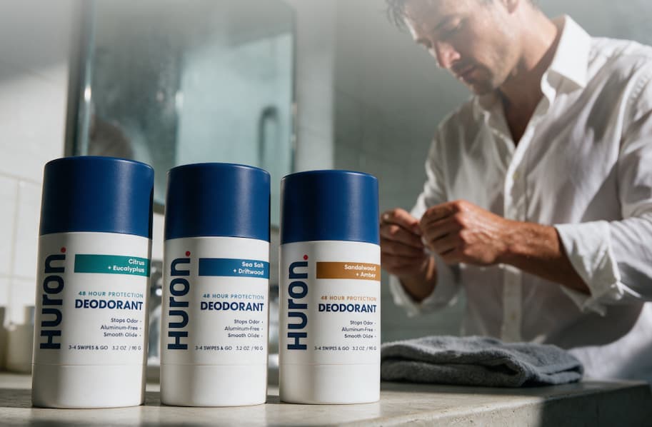 Three Huron deodorant sticks on bathroom counter, man getting ready in background