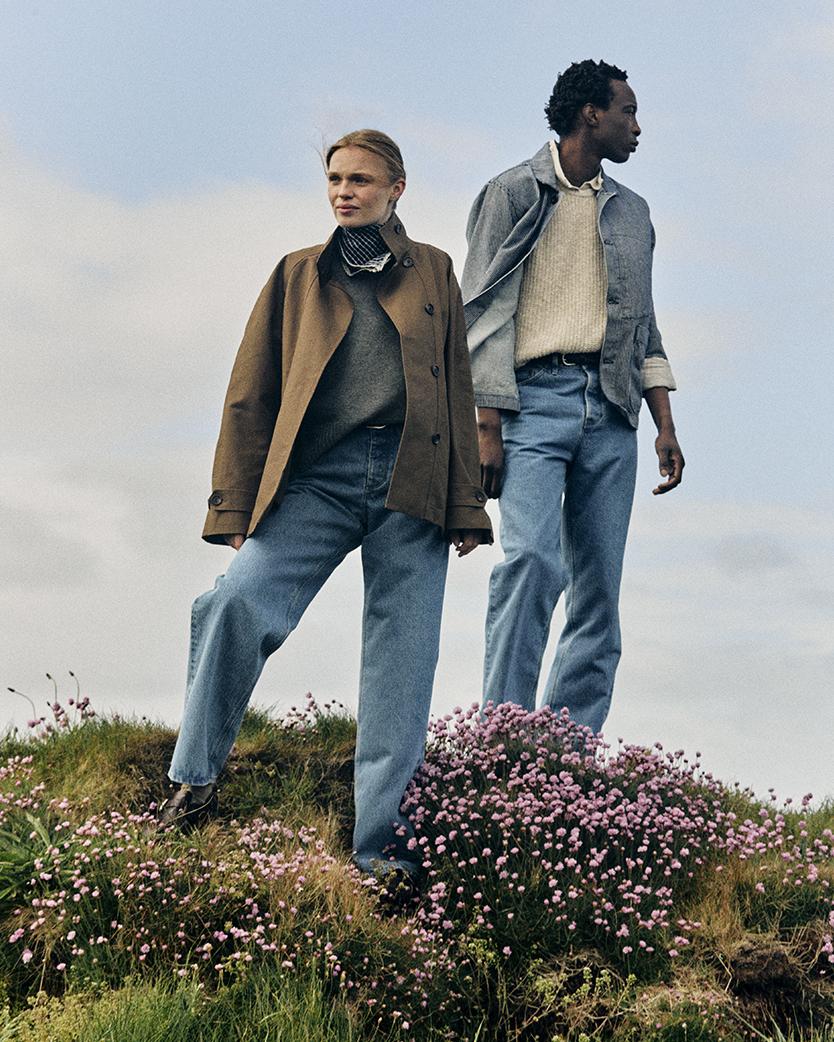 Man and woman stand side by side on grass and flowery mound. Both wear Summer Wash Selvedge denim jeans. She wears brown jacket over grey jumper. He wears grey shirt over cream wool jumper.