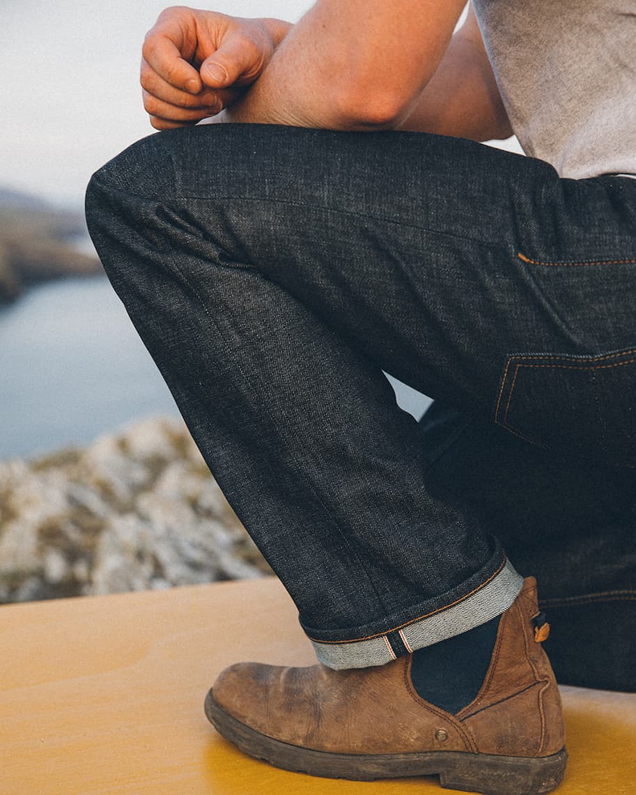 Image of leg bending down wearing selvedge denim jeans, and brown boots. Standing on yellow platform. Coast blurred into the background.
