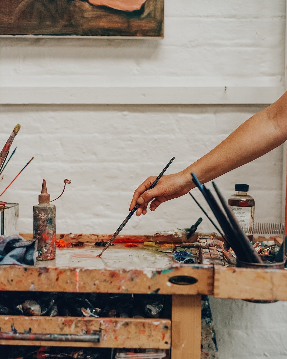 An arm holding a paintbrushes mixes paint on a palette full of paints and tools.