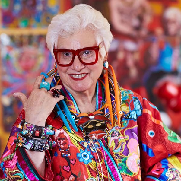 Portrait of Sue Kreitzman. She has white short hair, thick red glasses, wearing a colourful jacket.