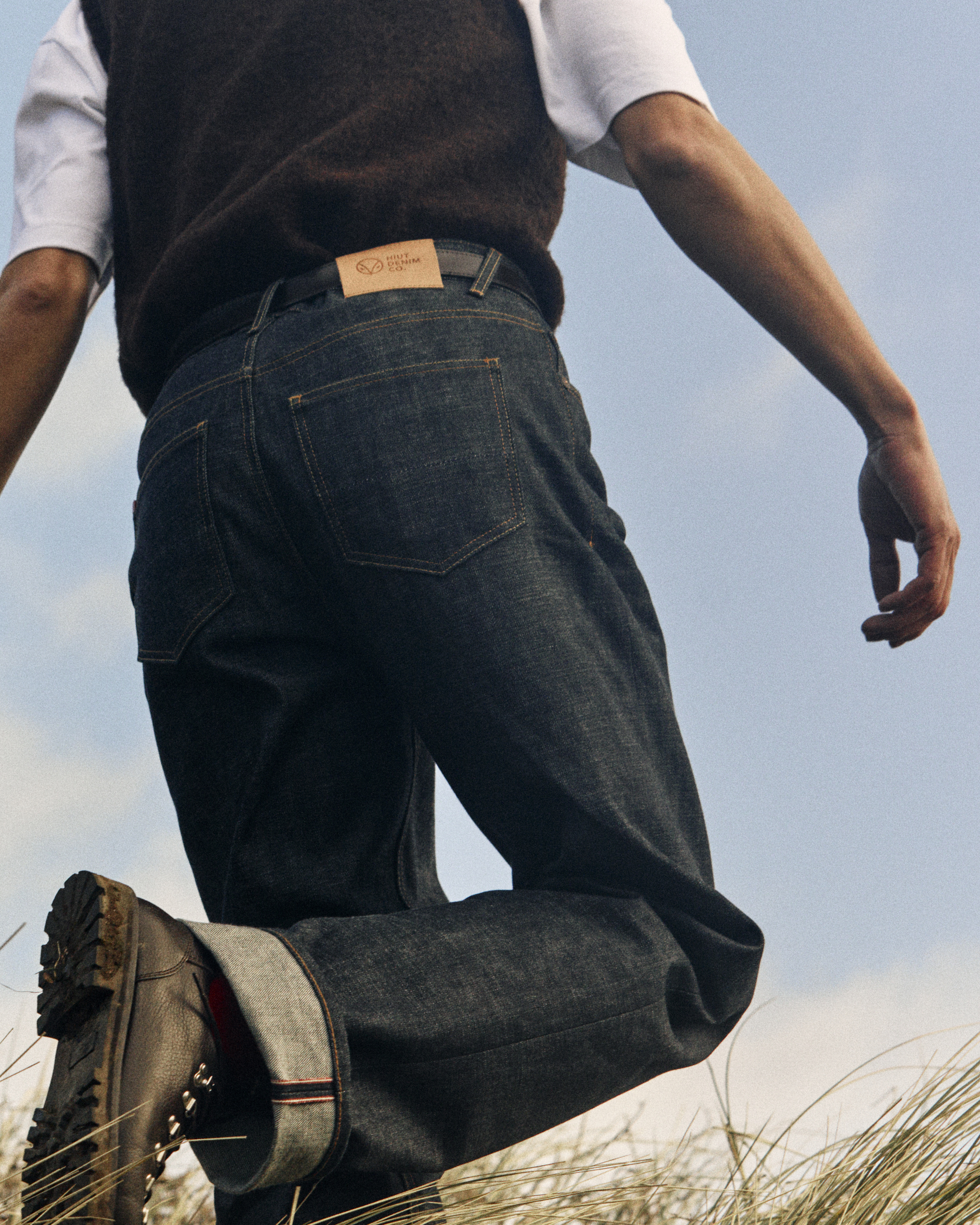 From behind image of man walking through dunes. He wears Miles selvedge denim Hiuts, white t-shirt, and wooden brown vest.