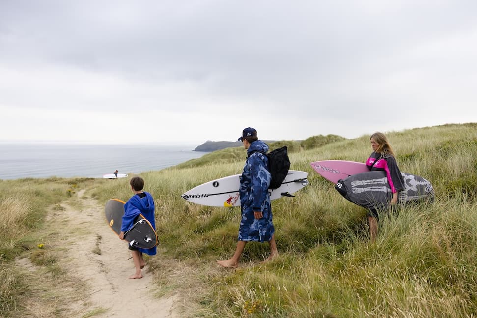 A family walking through the dunes towards the beach while carrying surfboards and wearing changing robes