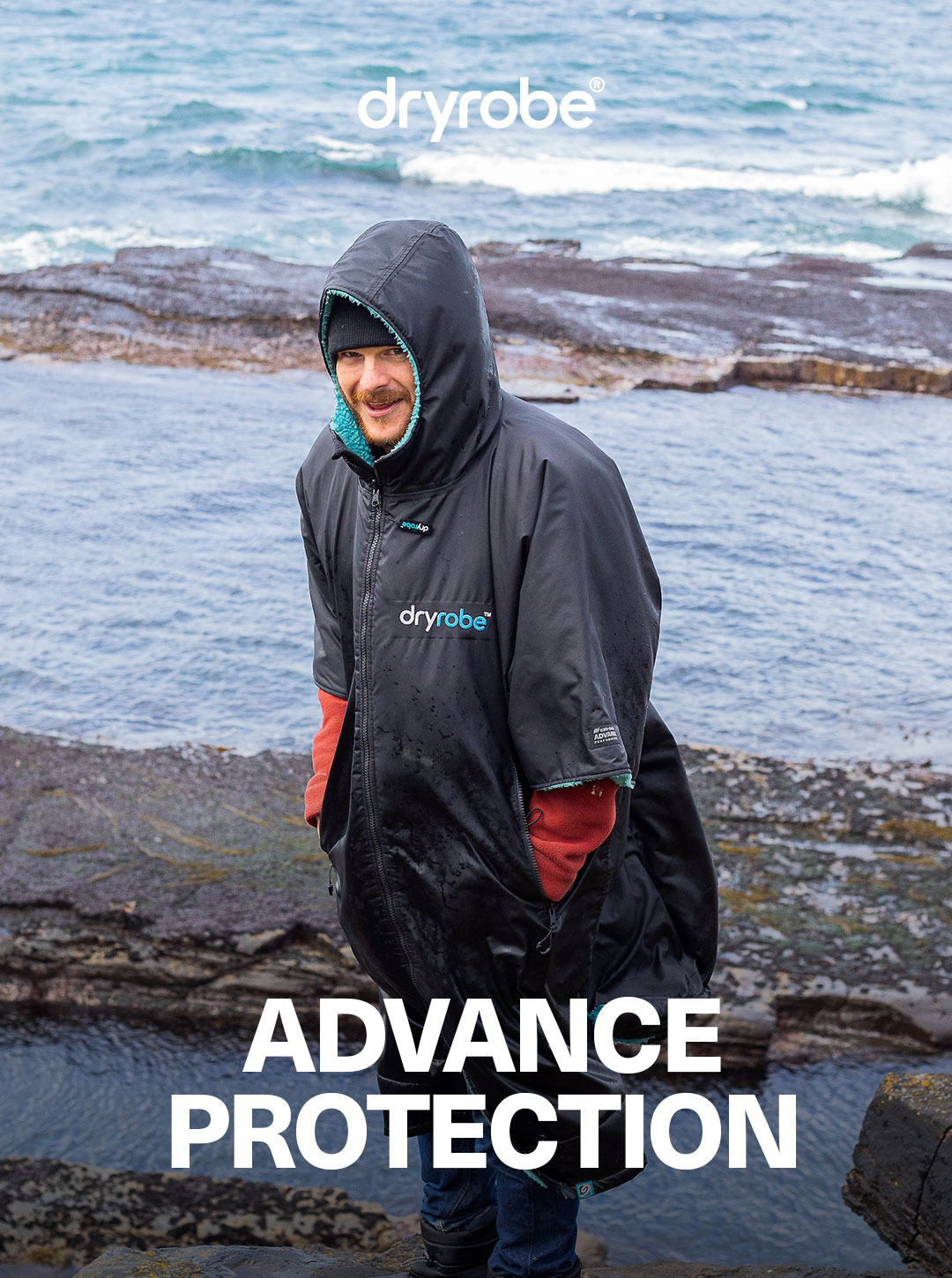 Man stood beside a rockpool and the sea while wearing a short sleeve Dryrobe to protect himself from the wind and rain