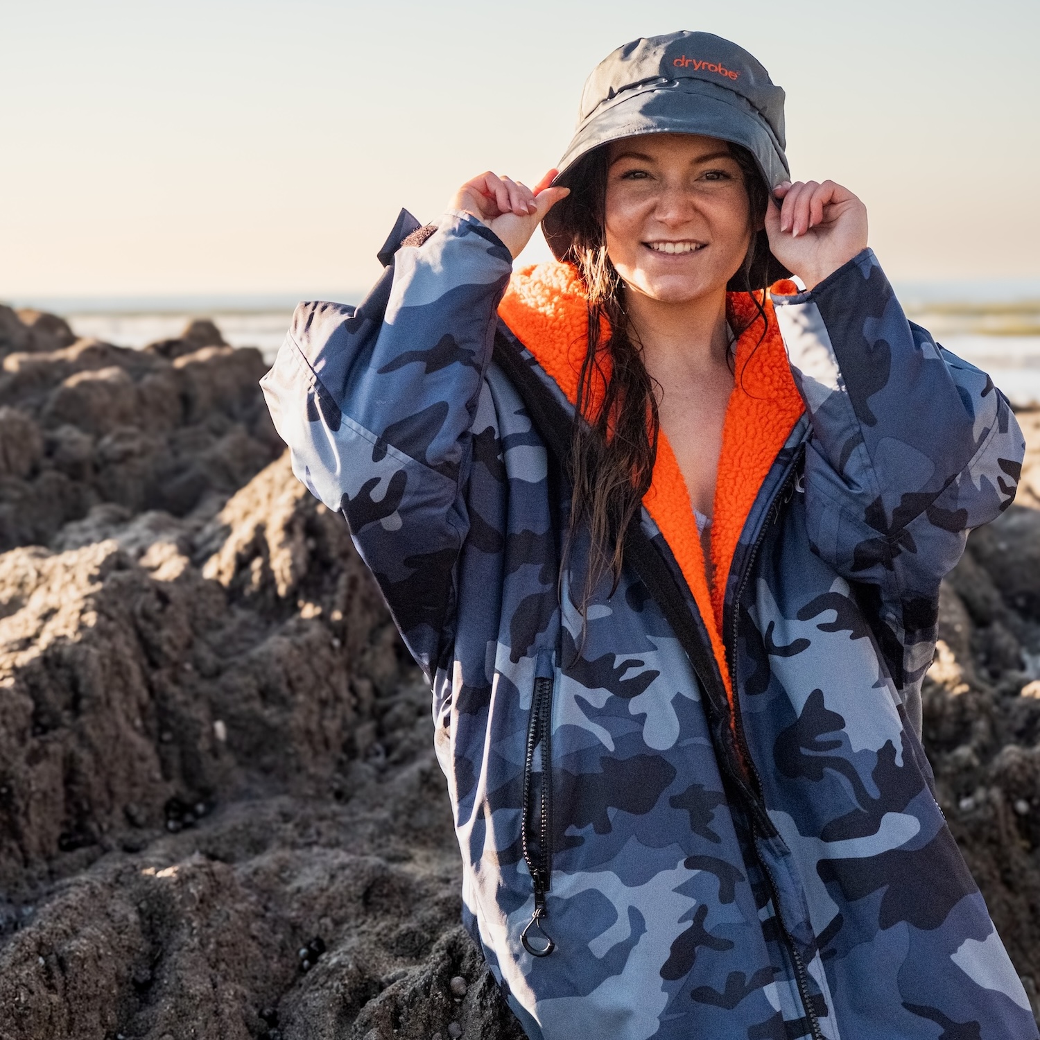 Smiling female wearing a bucket hat and dryrobe® Advance while sat on the rocks at the beach