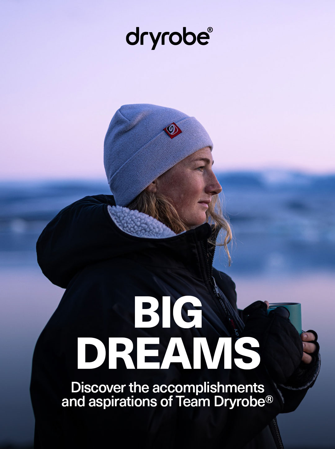 Woman wearing a Dryrobe® and beanie hugging a hot drink with snow covered mountains as a backdrop
