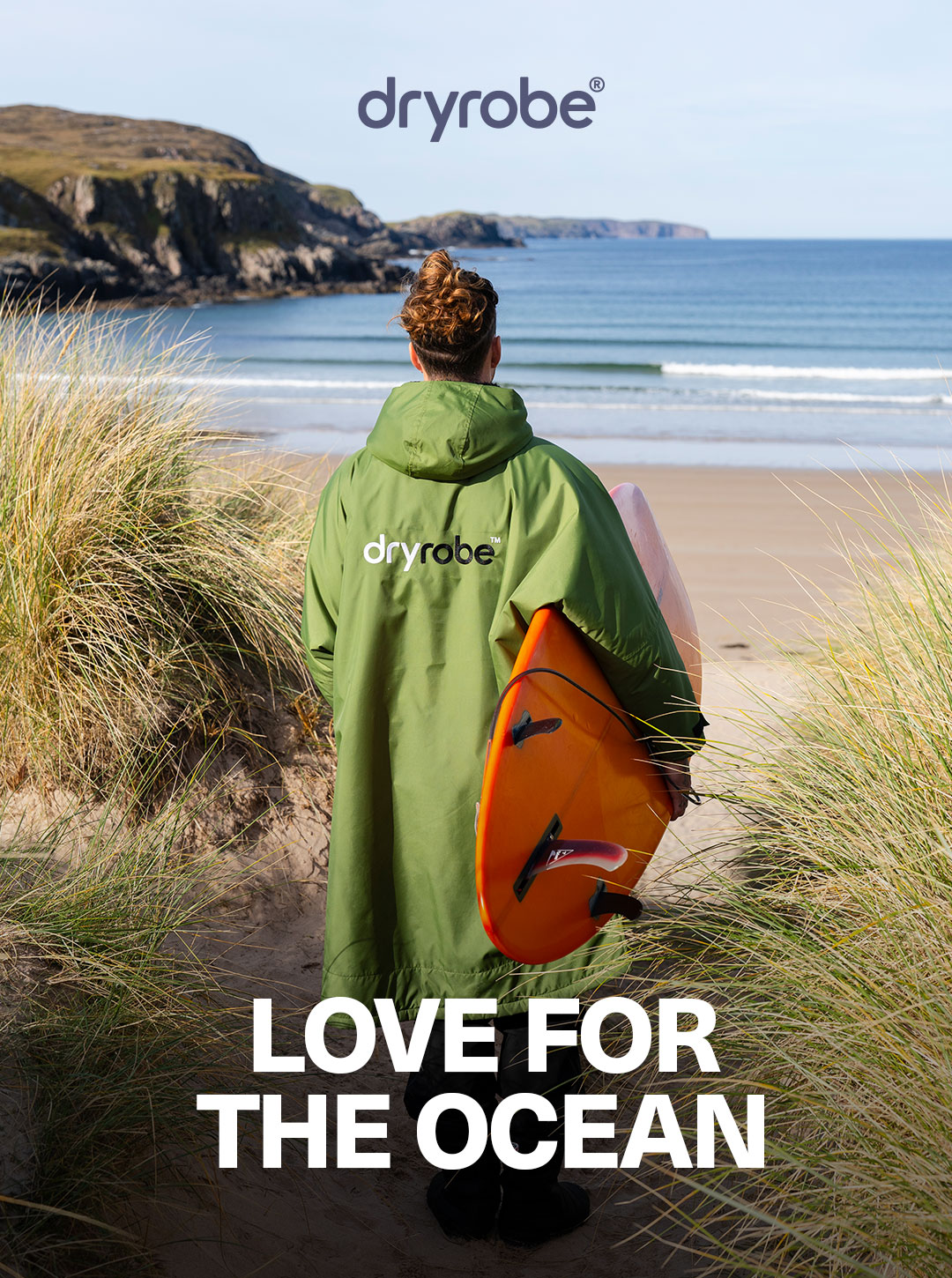 Male wearing dryrobe® gear looking out to sea on the sand dunes with a surfboard under his arm