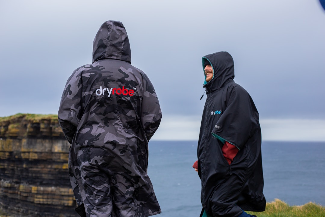 Two surfers stood on a cliff edge by the sea wearing Dryrobes