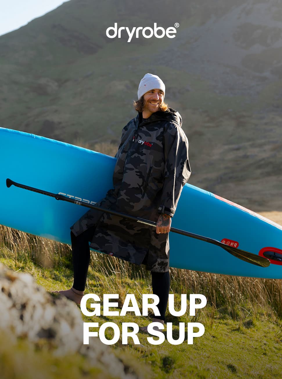 Man carrying a paddleboard up a grassy verge in the sunshine