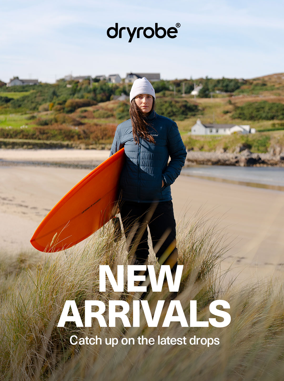 Woman wearing dryrobe® jacket and trousers while holding a surfboard with a backdrop of a beach