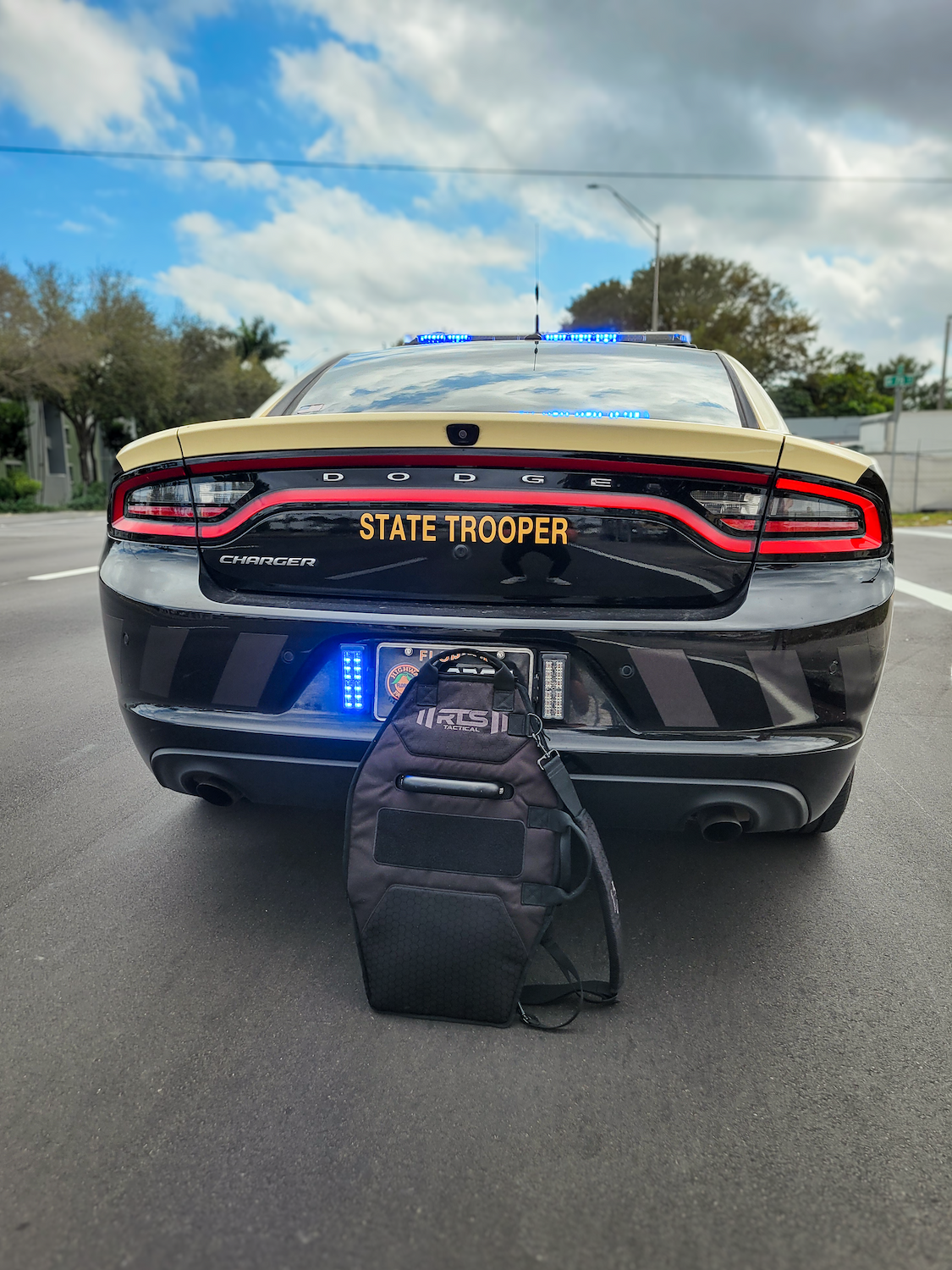 Rear view of a Dodge Charger state trooper patrol car with blue emergency lights activated, parked on a street, with an RTS Tactical black shield carrier bag placed behind the vehicle.