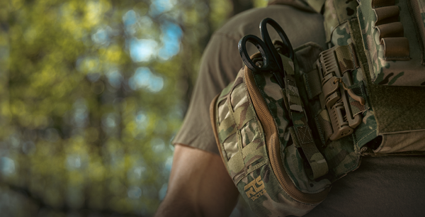 Close-up of a camouflage tactical vest with mounted medical shears in a MOLLE pouch, designed for quick-access gear deployment in the field