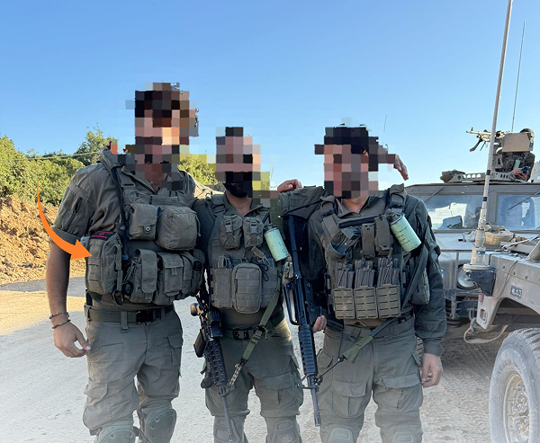 Three armed soldiers in tactical gear stand together outdoors in front of a military vehicle. Each is wearing plate carriers loaded with pouches and equipment. One soldier is marked with an orange arrow pointing to a IFAK on his waist.