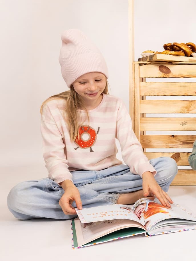 Sitzendes, lesendes Mädchen mit Pullover Doughnut und Mütze MALU