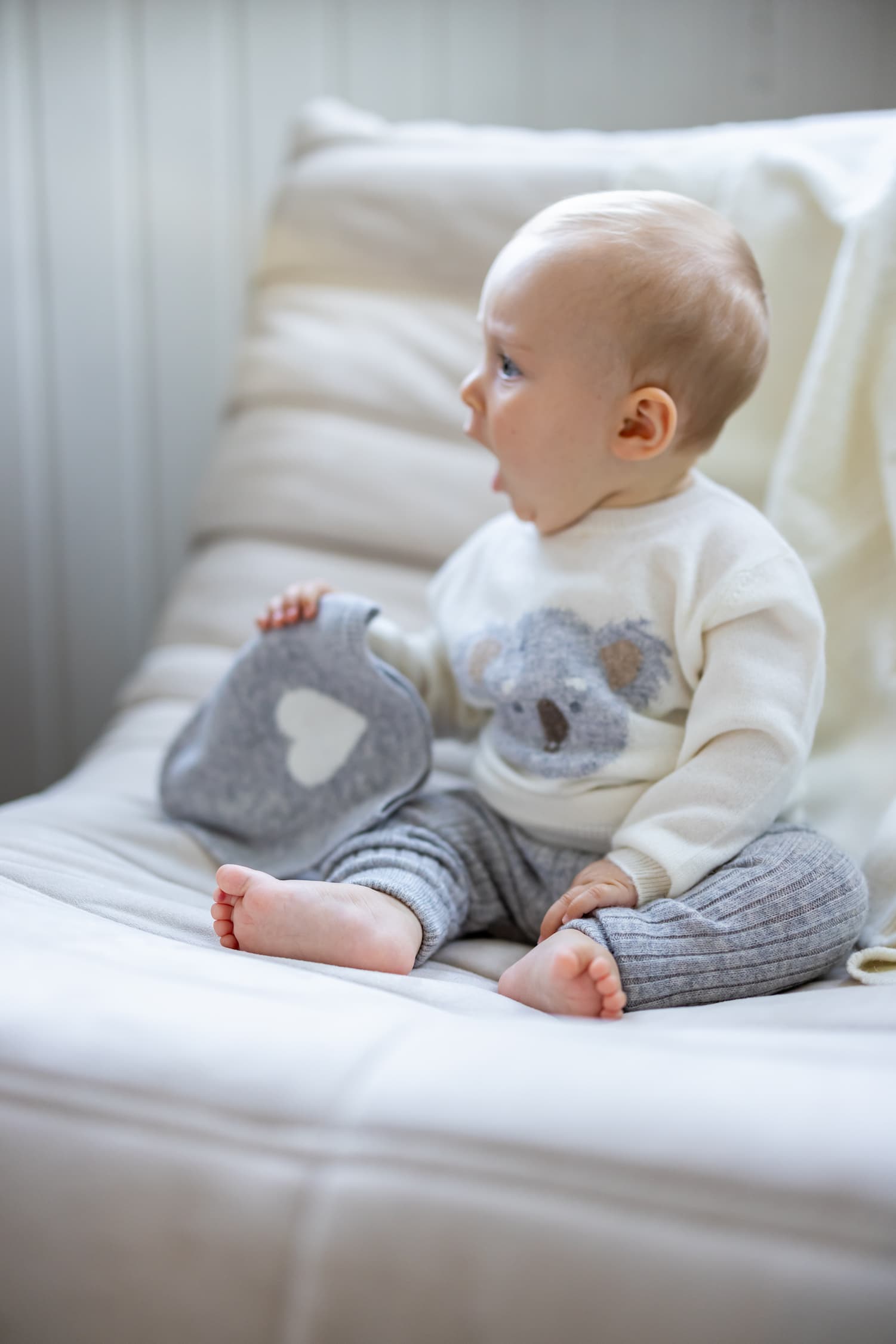 Baby sitzend auf Sofa mit Kaschmir Pullover KOALA hält hellgraue Kaschmirmütze HEART in der Hand