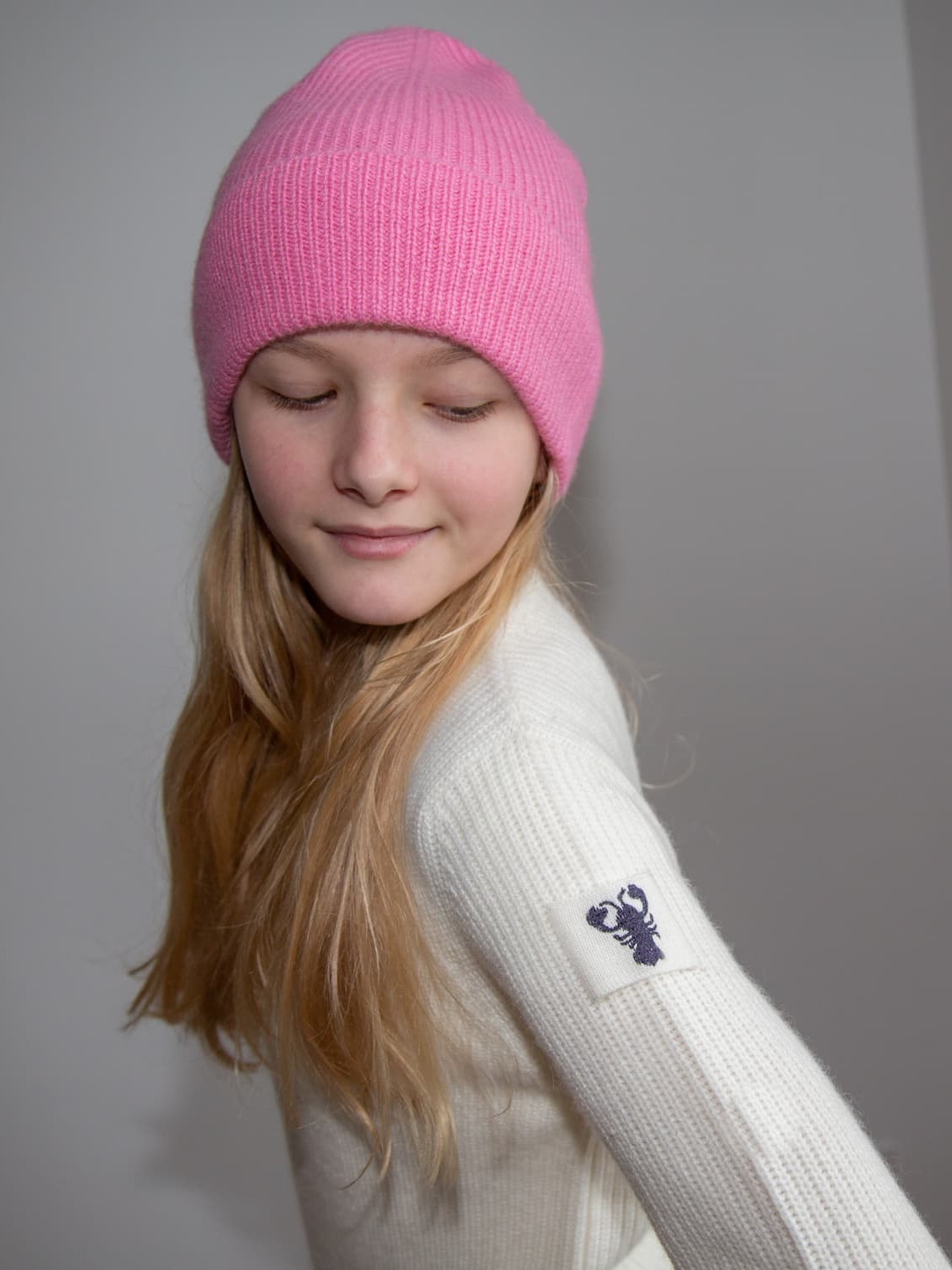 Bemütztes Mädchen mit weißem Pullover JENSON schaut auf Lobster-Emblem am Ärmel