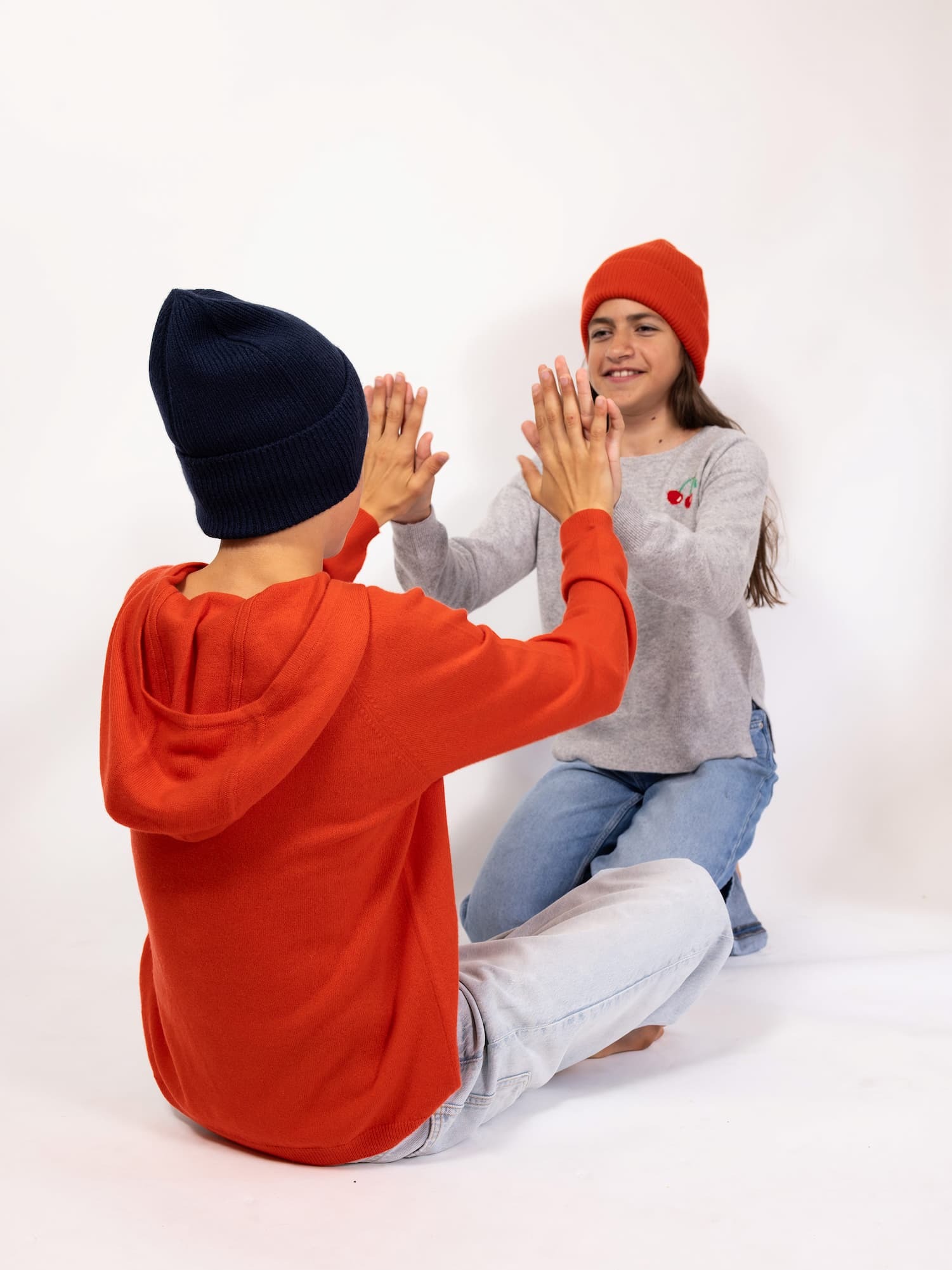 Teenie-Junge und -Mädchen sitzend mit Klatschspiel beschäftigt in Pullover CHERRY, Hoody JONA und Mütze MALU