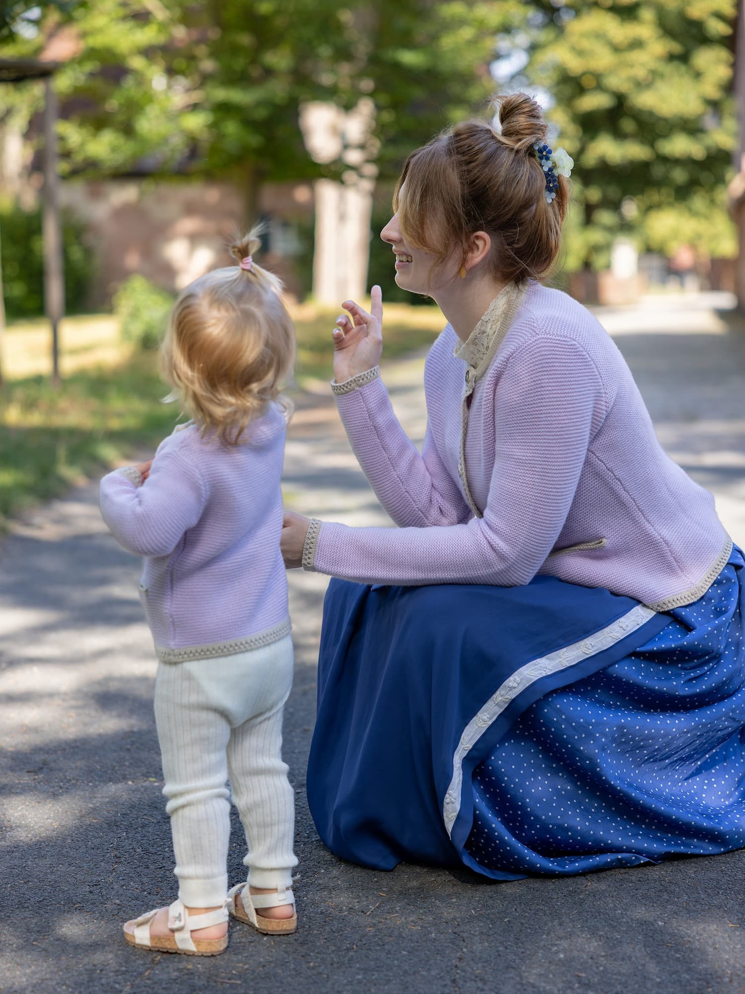 Hockende Frau neben stehendem Toddler-Girl mit Cardigan LENI in Mauve und mit Dirndl