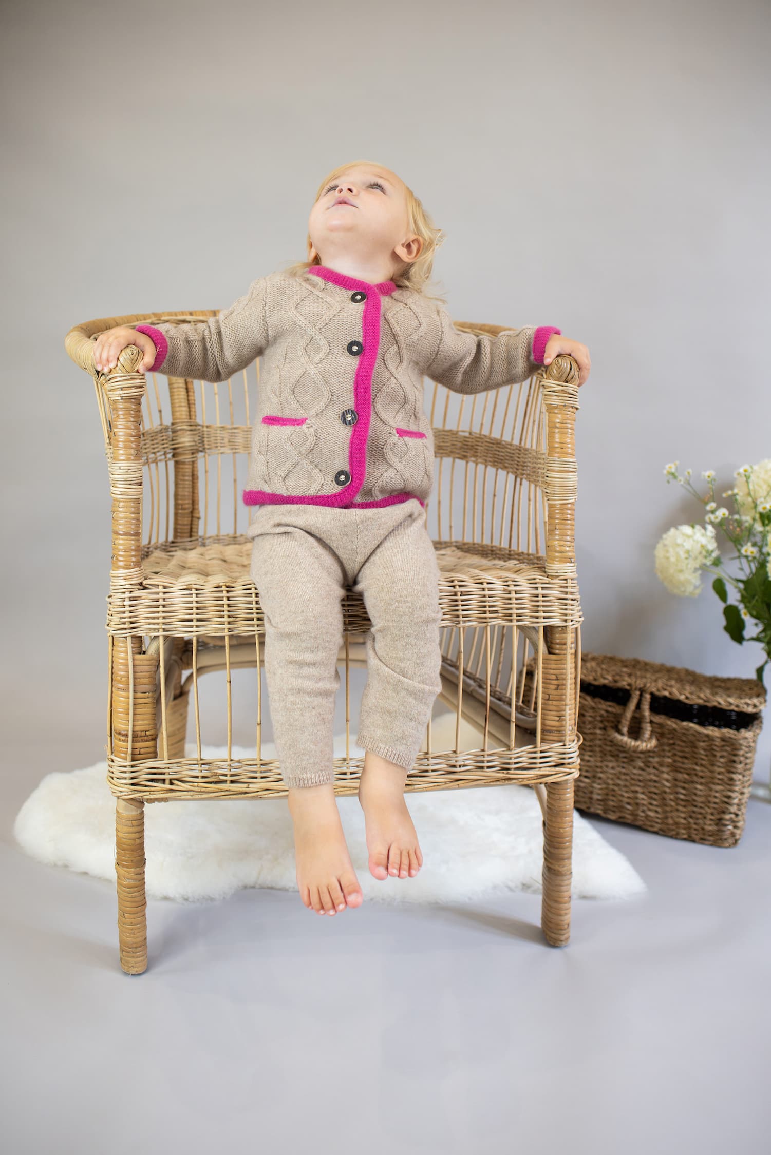 Toddler Mädchen in Kaschmirjacke MAXI sitzt auf Korbstuhl und schaut nach oben.