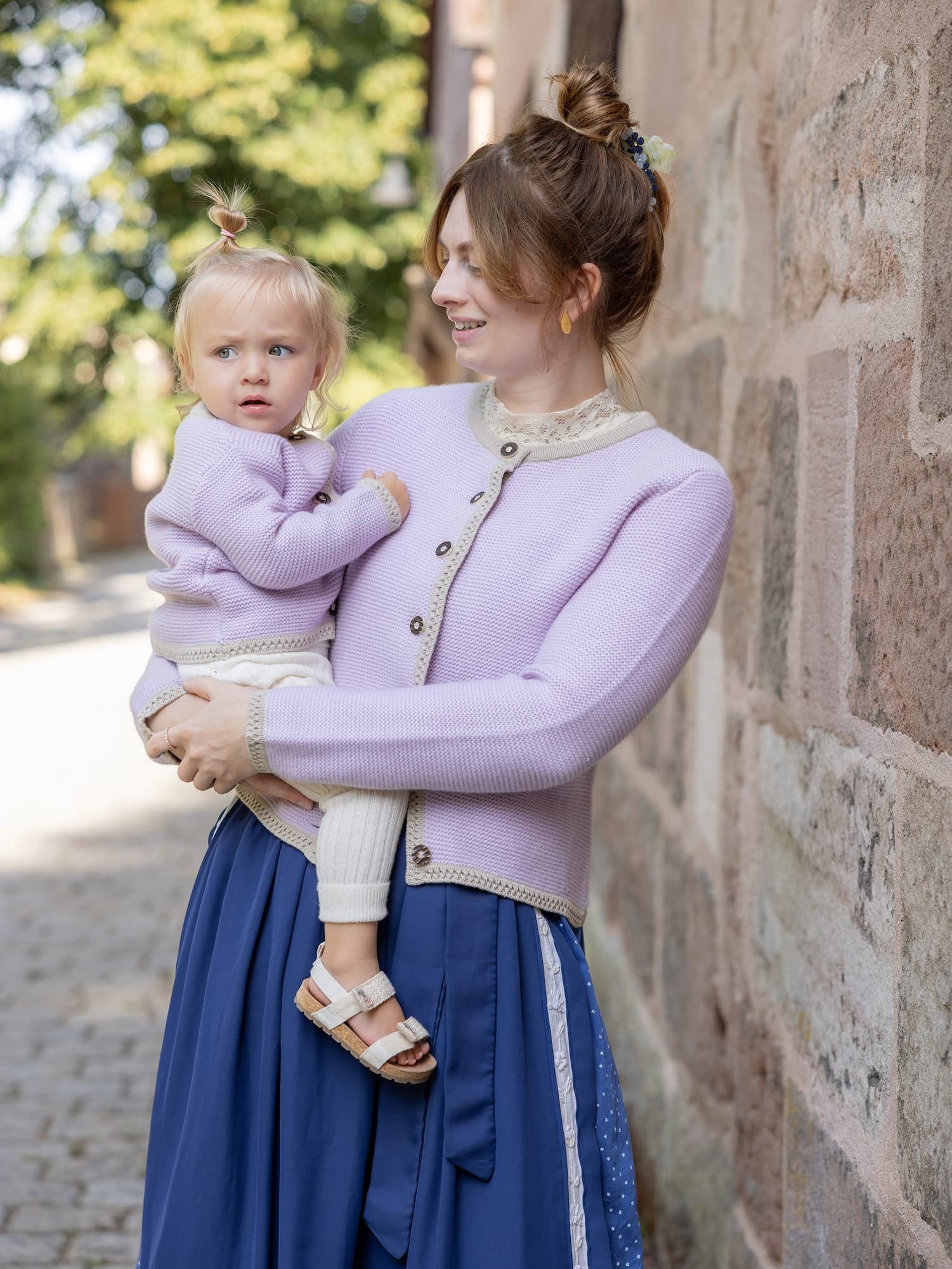 Stehende Frau mit Toddler-Girl im Arm, beide mit Cardigan LENI in Mauve und mit Dirndl